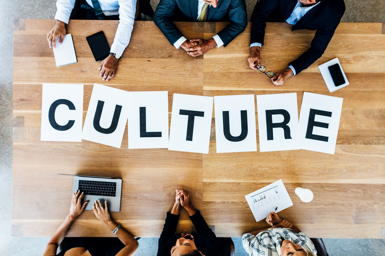 an overhead image of the word culture is spelled out in black uppercase letters on white pieces of paper laid out on a wooden table with six people at the table